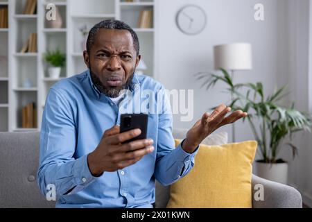 Middle-aged man sitting on couch, expressing confusion and disbelief while staring at his phone, looking at camera. Stock Photo