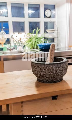 A mortar and pestle sits on a wooden counter in a kitchen. The kitchen is well-lit and has a modern feel Stock Photo