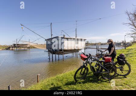 Woman cycles on an e-bike through the Po Delta in Northern Italy. In the background is a typical Italian pile dwelling, called Trabucco Stock Photo