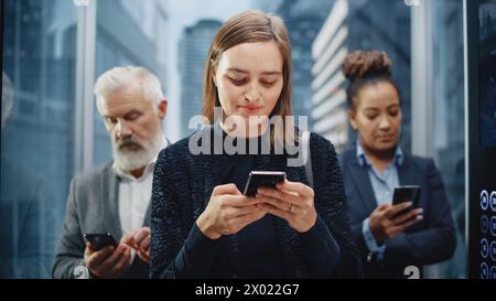 Three Diverse Multiethnic International People Ride a Glass Elevator to Office in a Modern Business Center. Focus on Young Confident Businesswoman Using Smartphone in a Lift. Stock Photo