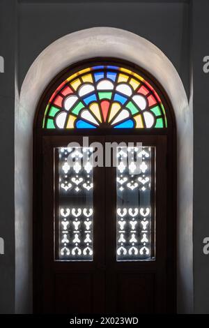 MUHARRAQ, BAHRAIN - FEBRUARY 10, 2018:  Light shines through stained glass and carved wooden shutters in the balcony doorway of a restored traditional Stock Photo