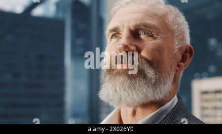 Portrait of Successful Middle Aged Businessman in a Perfect Suit Standing in Modern Office Looking out of the Window on Big City with Skyscrapers. Successful Finance Manager with Great Career. Stock Photo