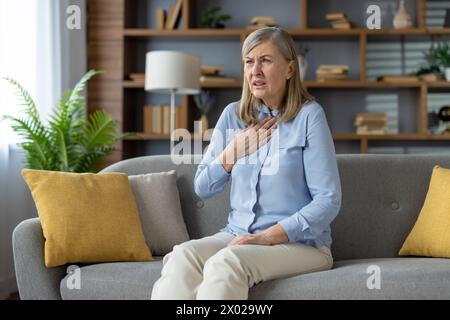 Senior woman touching chest while having pain in throat after coughing in domestic interior. Disturbed lady in white pants feel sick from virus or bacteria infection and having loss of voice. Stock Photo