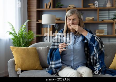 Sad elderly lady sitting on pillowed sofa under blanket and getting ready for taking medicine with glass of water. Struggling woman suffering from headache and using painkillers during sick leave. Stock Photo