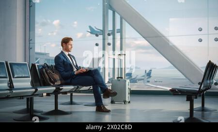 Modern Airport Terminal: Handsome Businessman Working on Laptop Computer While Waiting for His Flight. Man Sitting in a Boarding Lounge of Big Airline Hub with Airplanes Departing and Arriving Stock Photo