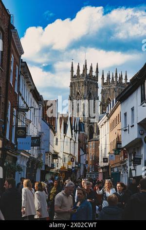 Bustling street scene with pedestrians in a historic city center, featuring old buildings and a prominent Gothic cathedral under a cloudy sky in York, Stock Photo