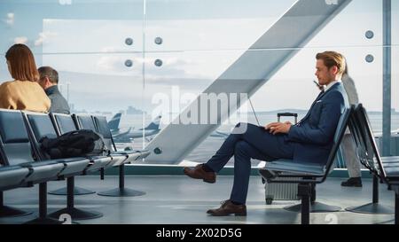 Airport Terminal: Businessman Uses Laptop Computer, Waiting for Flight to International Conference. Traveling Entrepreneur Remote Work Online Sitting in a Boarding Lounge of Airline Hub with Airplanes Stock Photo