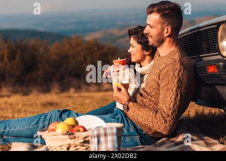 Beautiful young couple enjoying picnic time on the sunset. They drinking tea and sitting in a meadow leaning against a old fashioned car. Stock Photo