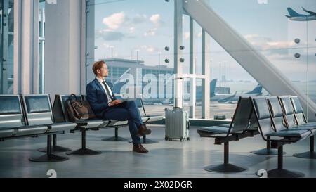 Airport Terminal: Businessman Uses Laptop Computer, Waiting for His Flight. Traveling Entrepreneur Remote Work Online Sitting in Boarding Lounge of Airline Hub with Airplanes Arriving, Departing. Wide Stock Photo