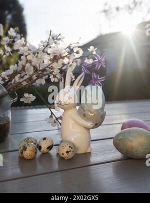 souvenir porcelain Easter bunny stands on the table in the rays of the setting back sun and holds an egg. spring white blossoming tree branches. Home Stock Photo