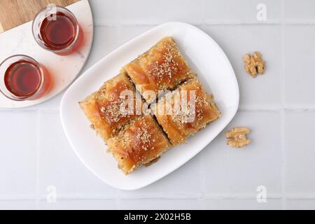 Eastern sweets. Pieces of tasty baklava, walnuts and tea on white tiled table, flat lay Stock Photo