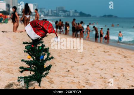 A makeshift small traditional Christmas tree and Santa Claus hat planted on the sand at Leblon Beach with a defocused crowd in the background Stock Photo