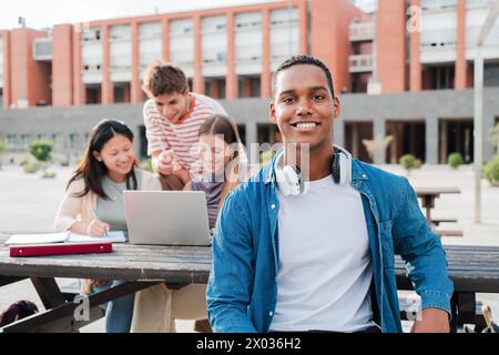 Young african american male student, smiling and looking at camera sitting with her classmates at university campus. Schoolboy staring front behind her multiracial friends while they use a laptop. High quality photo Stock Photo