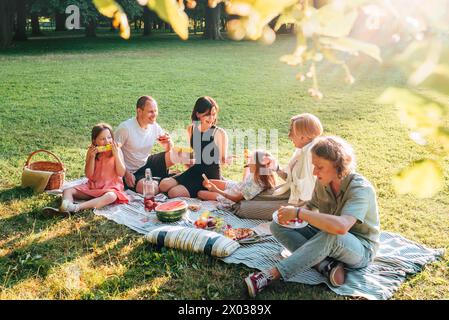 Big family sitting on the picnic blanket in city park during weekend Sunday sunny day. They are smiling, laughing and eating boiled corn and watermelo Stock Photo