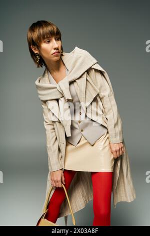 A chic young woman with short hair, donning tights and a coat, holds a fashionable handbag in a studio setting. Stock Photo