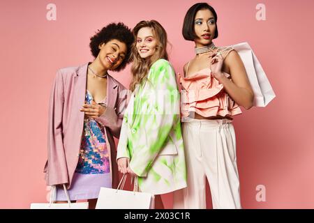 Three women from diverse backgrounds stand together, holding colorful shopping bags in front of a pink studio background. Stock Photo