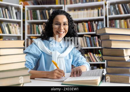 Cheerful Hispanic female student with glasses studying diligently among towering stacks of books in a library. Stock Photo