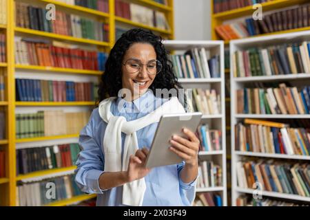 Smiling young woman in glasses using a digital tablet among bookshelves in a library, representing modern learning and technology in education. Stock Photo