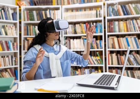 Young woman wears virtual reality headset in library, experiencing immersive technology with books and laptop around. Stock Photo