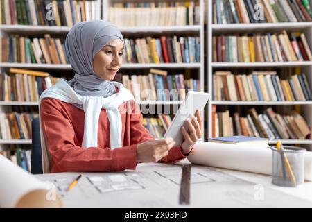 A focused Muslim woman in a hijab using a digital tablet at a library table with architectural blueprints and books around. Stock Photo