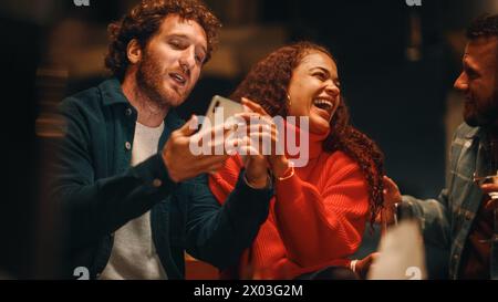 Man and Woman Sitting on a Date at an Outdoors Restaurant Terrace on a Warm Summer Evening. Loving Mixed Race Couple Using Smartphone, Looking on Photos, Checking Social Media, Having Fun. Stock Photo