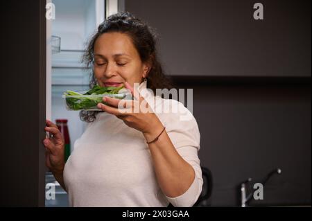 Young woman pulls out a recyclable cardboard crate with ecologically friendly organic green spinach leaves from the refrigerator, enjoying the aroma o Stock Photo