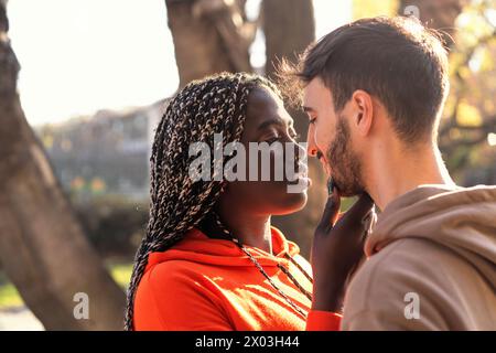 A young African woman with braided hair gently touches the face of a man - gazes locked in a tender embrace. Multiracial lovers at sunset - diverse lo Stock Photo