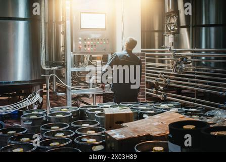 Brewmaster monitors beer production in craft brewery amidst kegs and shiny stainless steel barrels Stock Photo