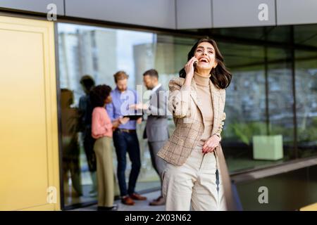 A stylish woman converses on her smartphone, walking past colleagues gathered in casual conversation Stock Photo