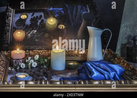 A still life with a blue satin fabric next to several lit candles, flowers surrounded by a golden frame and their reflection on a black surface Stock Photo