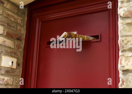 A newspaper is halfway through a mail slot in a red door framed by bricks, indicating a recent delivery. Stock Photo