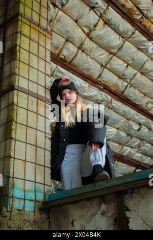 Beautiful teenage girl in a black winter jacket and hat poses in destroyed and abandoned building. Urban concept. Vertical shot. Stock Photo