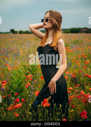 Beautiful young girl in a black evening dress and sunglasses posing against a poppy field on a cloudy summer day. Portrait of a female model outdoors. Stock Photo