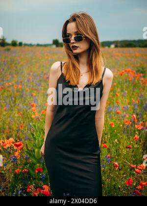 Beautiful young girl in a black evening dress and sunglasses posing against a poppy field on a cloudy summer day. Portrait of a female model outdoors. Stock Photo