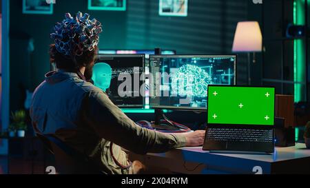 IT admin coding and using EEG headset to upload brain into green screen laptop, gaining immortality. Neuroscientist engineering transhumanism experiment on chroma key device, camera A Stock Photo
