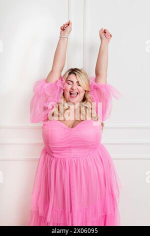 Enthusiastic plus size female in stylish outfit standing and raising arms behind white background Stock Photo