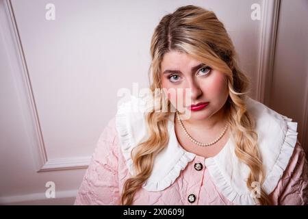 Stylish plus size woman in upset mood wearing bead necklace looking at camera against pink background at studio Stock Photo