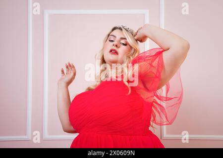Optimistic curvy woman in red dress with arm raised against head while dancing behind pink background Stock Photo