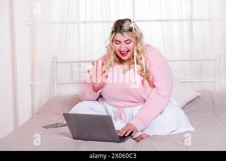 Positive curvy lady in sweater sitting on comfortable bed waving hand while using laptop to do video call in cozy room Stock Photo