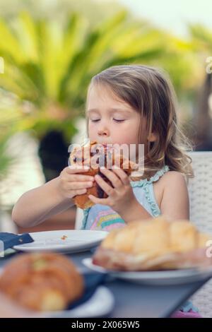 A beautiful little girl eats a croissant with chocolate with appetite. The girl has breakfast with a croissant. Caucasian girl eats pastries. Close-up Stock Photo