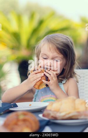 A beautiful little girl eats a croissant with chocolate with appetite. The girl has breakfast with a croissant. Caucasian girl eats pastries. Close-up Stock Photo