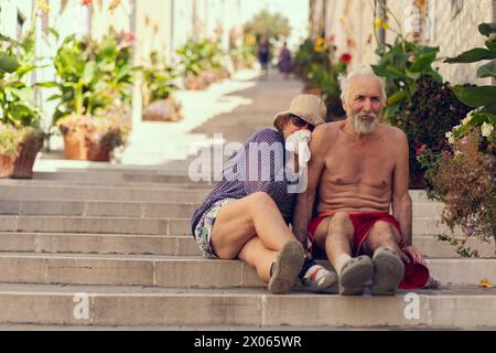 Old travelers are sitting on the steps of a long staircase. A long street with steps is lined with potted plants, an elderly couple smiles while sitti Stock Photo