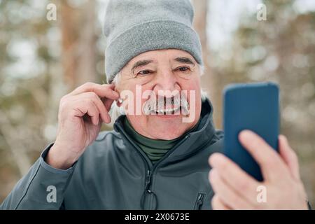 Medium closeup of cheerful senior man fixing wireless earphone while talking on video call Stock Photo