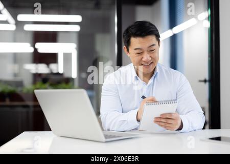 A cheerful Asian businessman is jotting down notes in a spiral notebook while working on a laptop in a contemporary office space. Stock Photo