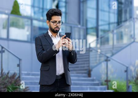 A young Indian businessman in a suit is standing outside an office building and using a mobile phone, typing, reading a message. Stock Photo