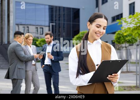 Businesswoman with four colleagues outside office building, company employee checking documents, smiling contentedly. Stock Photo