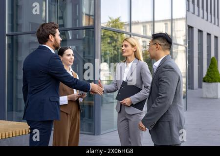 Interracial group of young business people standing on street outside office building in suits, man and woman shaking hands, making deal and getting to know partners and clients. Stock Photo