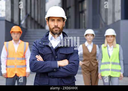 Portrait of serious young male engineer, owner in uniform and hard hat standing in front of camera with arms crossed on chest, behind is an interracial group of colleagues and contractors. Stock Photo