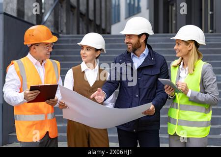 An interracial team of men and women, engineers and developers, stand in hard hats outside the building, holding a plan in their hands, discussing the project among themselves. Stock Photo
