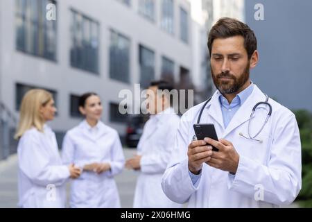 A serious young male doctor, scientist is standing outside a hospital room and is concentrating on using a mobile phone, colleagues are standing behind him and are looking at each other. Stock Photo